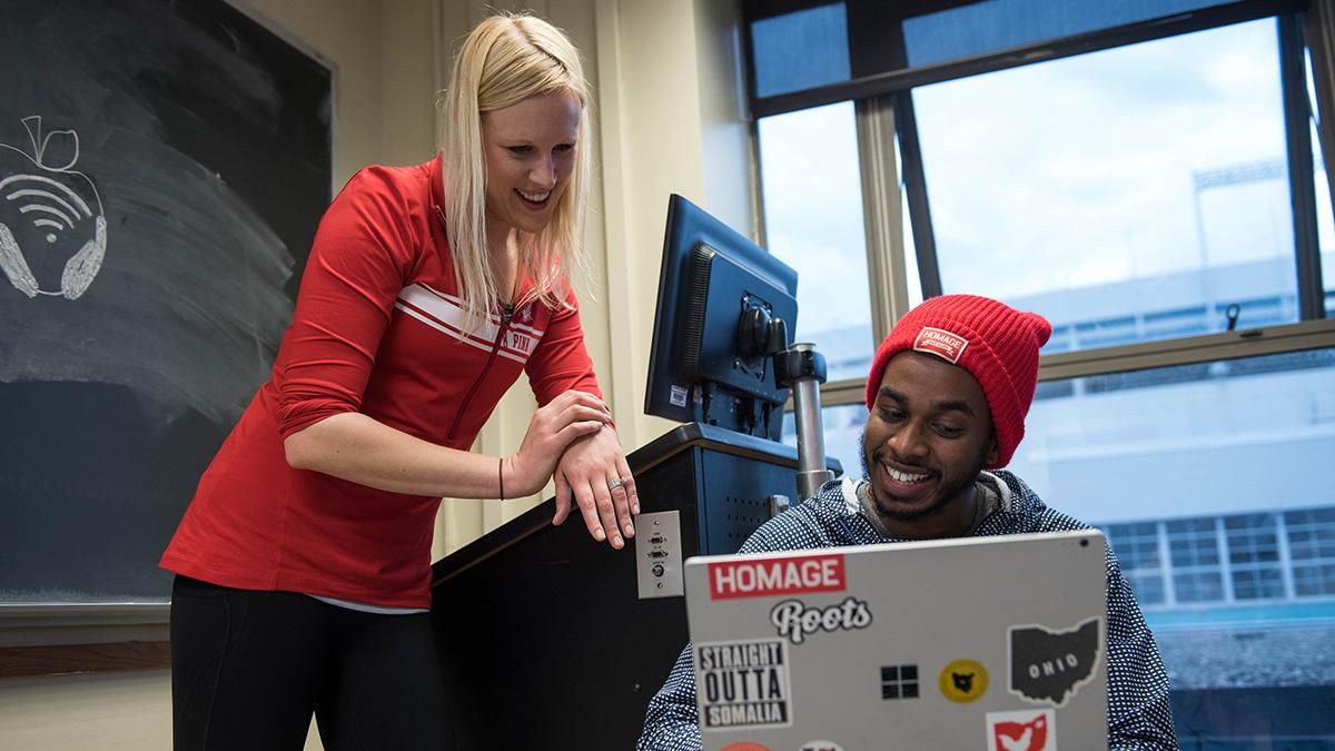 A student and faculty member talk while the student looks at his laptop.