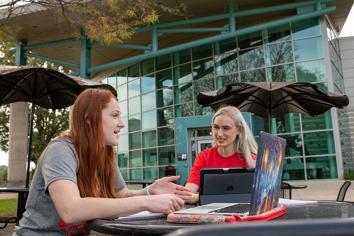 Two students sit outside a building on the Newark campus and talk while sitting in front of computers