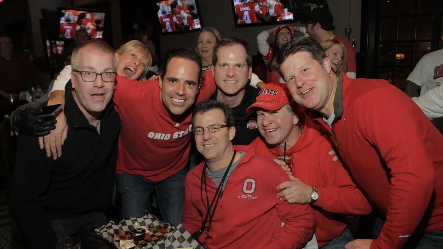 a group of alumni wearing ohio state gear pose for a group shot at a bar during an ohio state football game