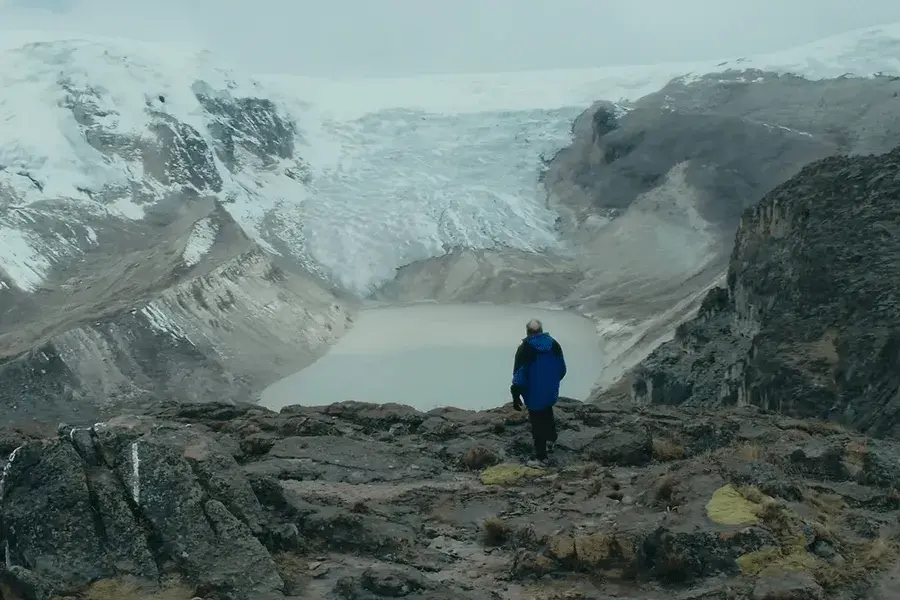 A man standing on the edge of a mountain overlooking a huge glacier and snow-capped mountains in the distance
