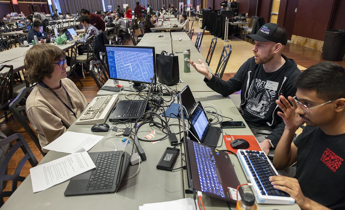 A group of students talk at a table with laptops and other technological devices in front of them