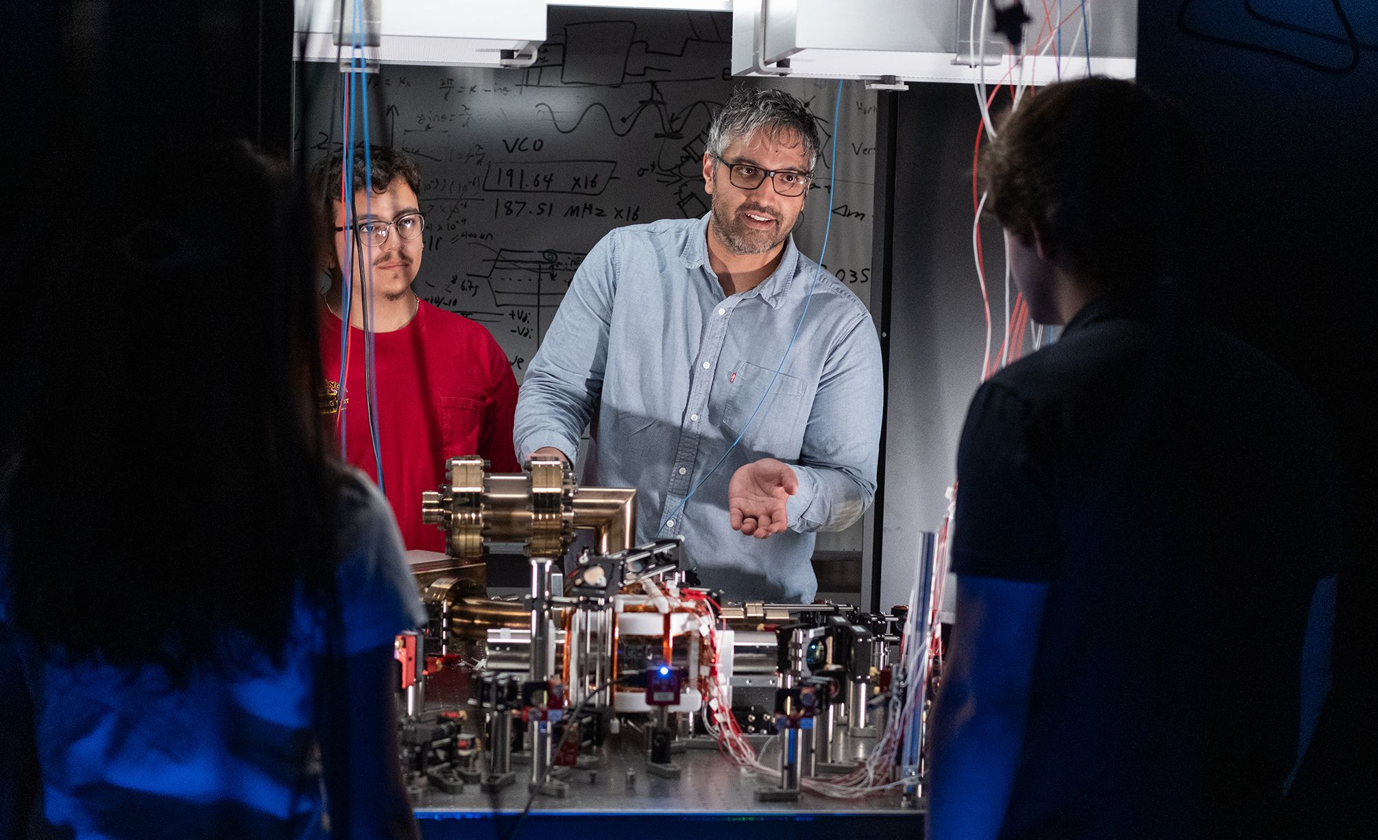 A researcher standing in front of a circuit board talks to colleagues