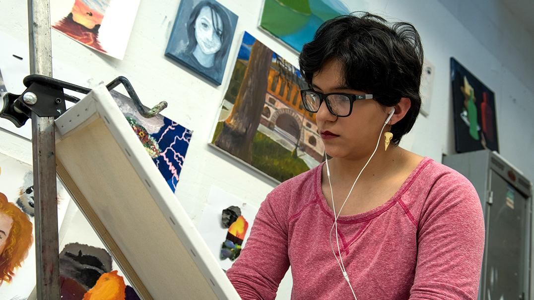 A student wearing headphones paints on a canvas in front of a wall full of hanging art