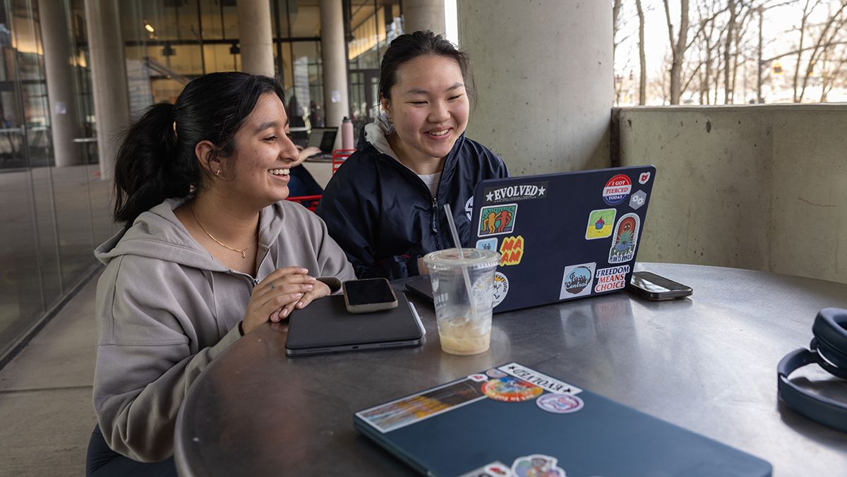 Two students sit in front of a laptop at a table.