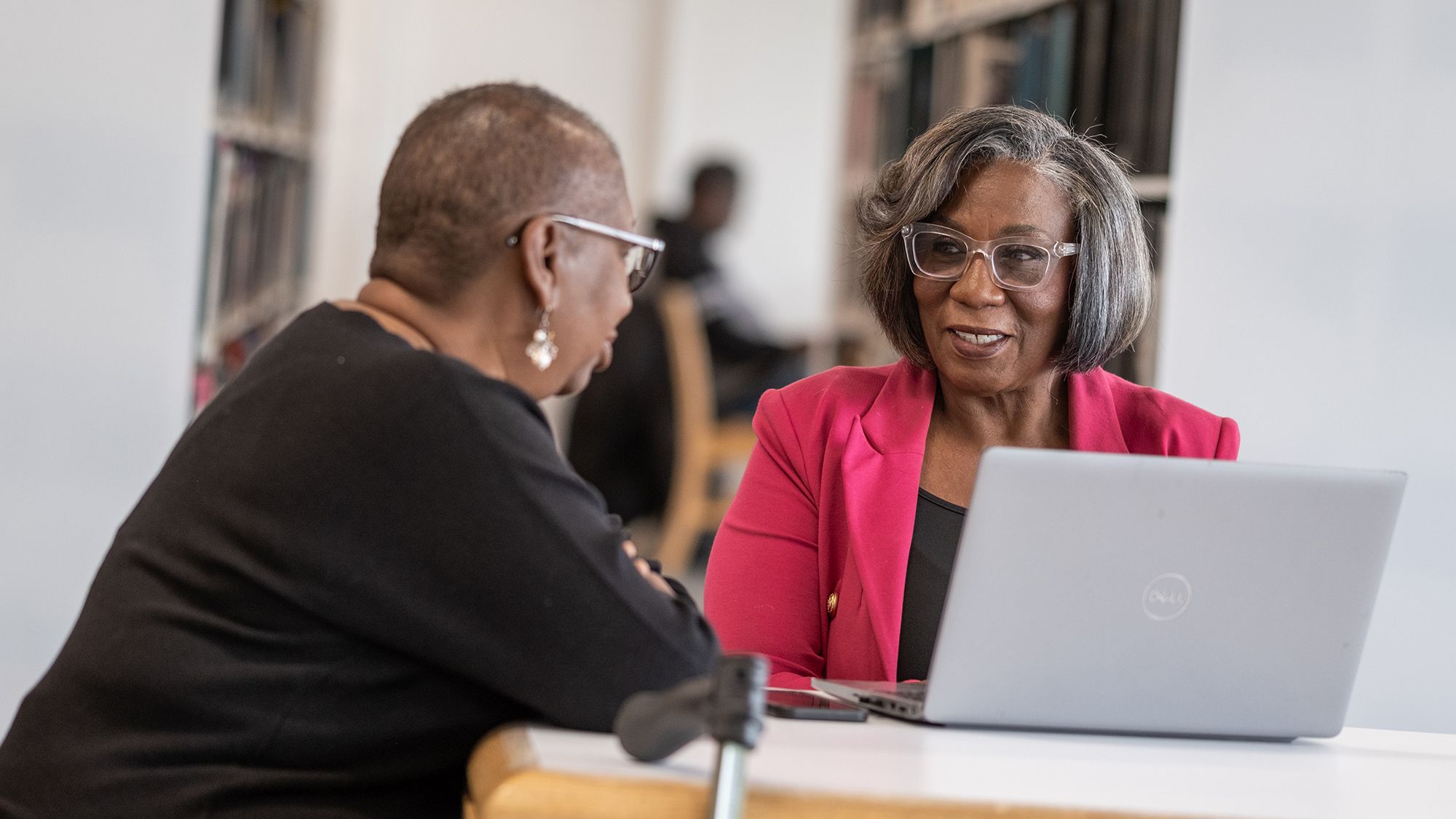 Two woman talk at a table while one sits in front of a laptop computer