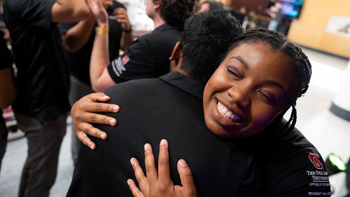 A close up of two students embracing each other in a hug. One of the students is smiling with their eyes closed while facing the camera