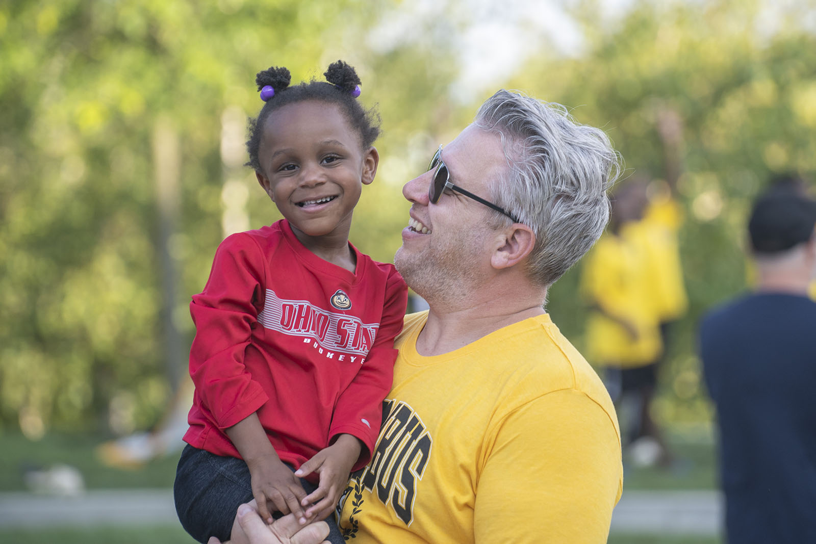 a father wearing a columbus crew T-shirt holds his smiling daughter who is wearing an Ohio State T-shirt