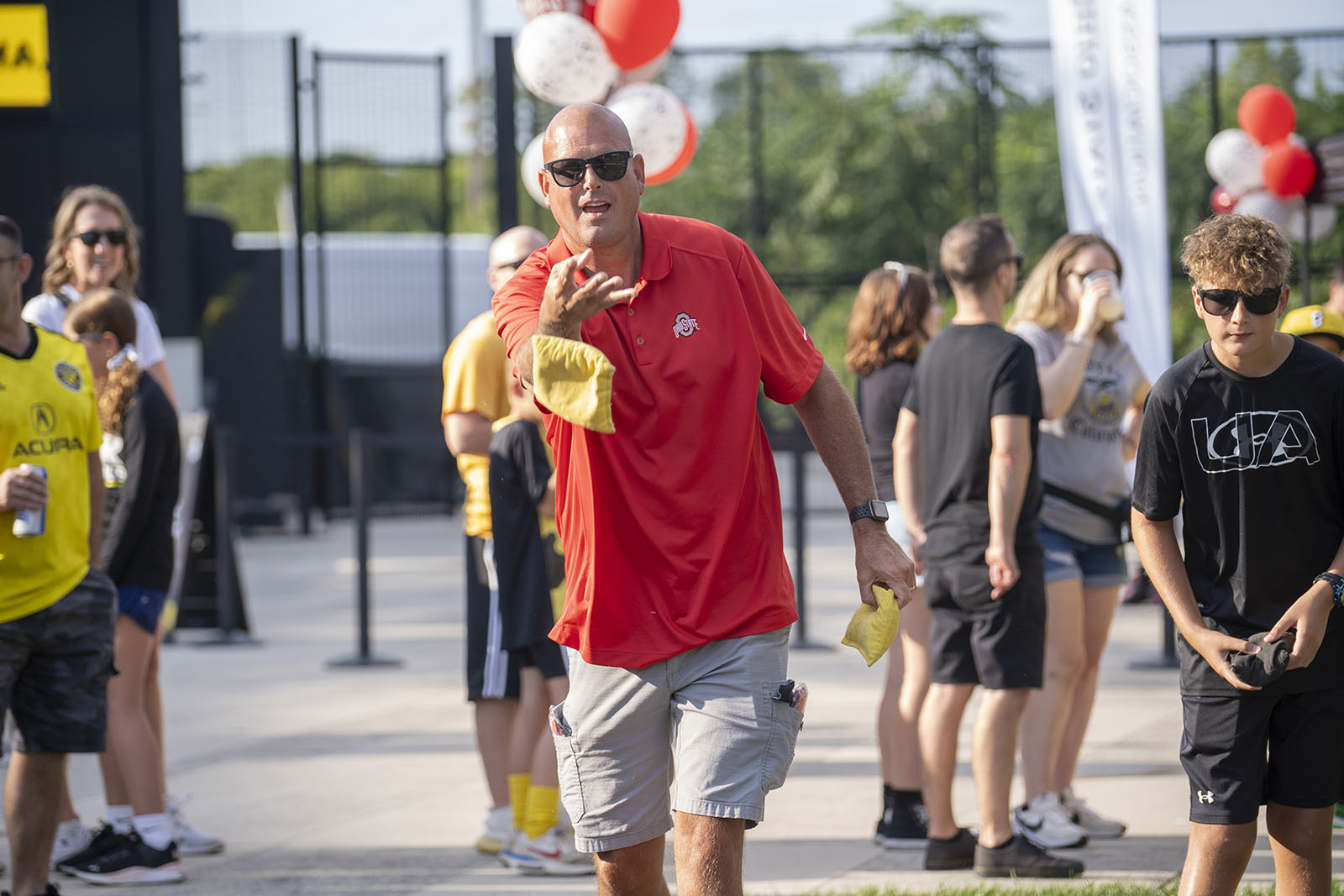 Person wearing a red shirt and black sunglasses tosses a bean bag