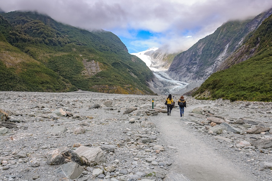 Two people walking on a narrow path lined with rocks and hills