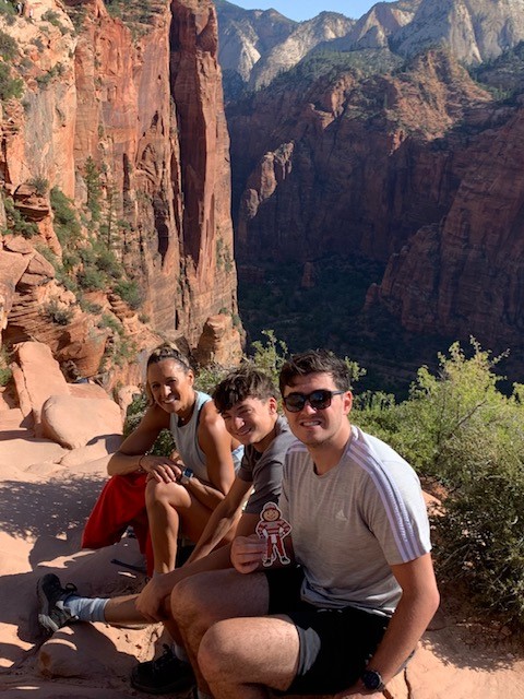 Brutus sitting with a woman and two men, along a cliff among red, rock mountains in Springdale, Utah.