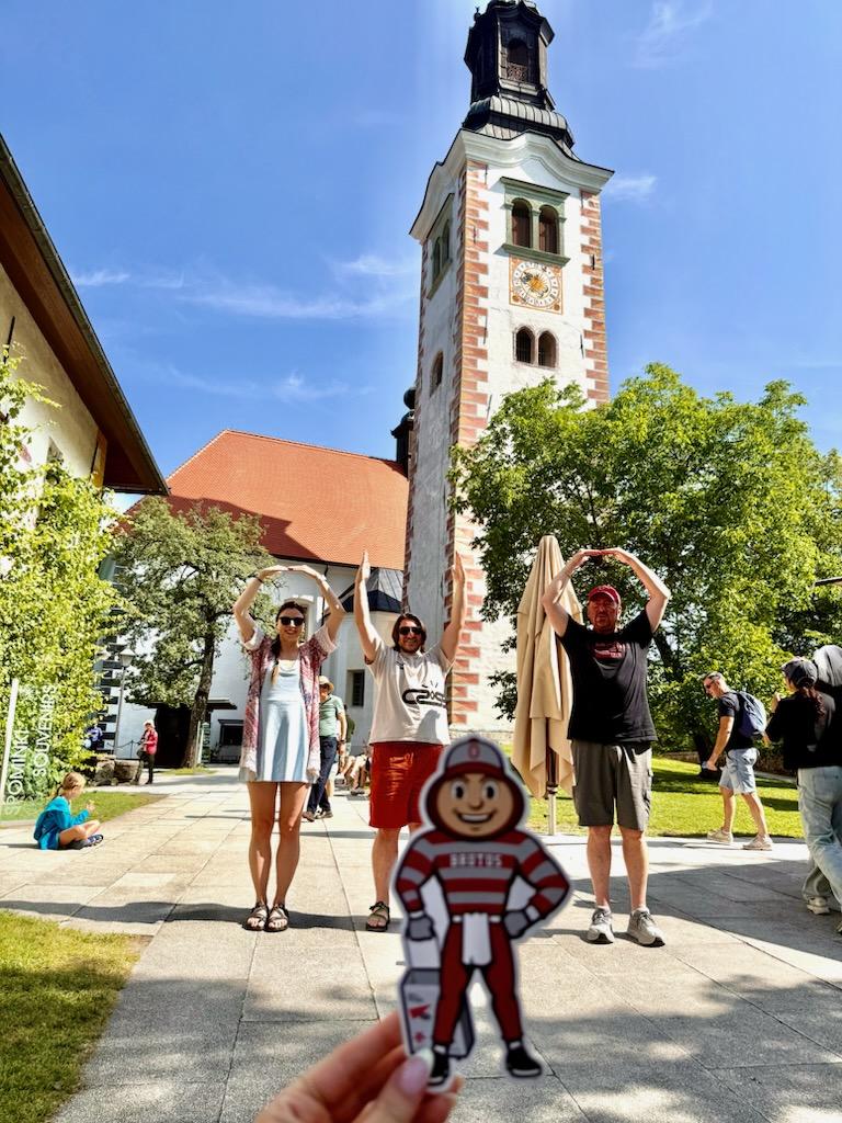 Brutus being the "I" in O-H-I-O of one women and two men in front of a church in Lake Bled, Slovenia.