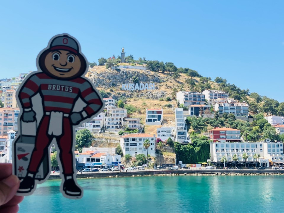 Brutus in front of blue water and a small resort town on a hill; halfway up the hill, a sign says "Kuşadası" in Turkey.