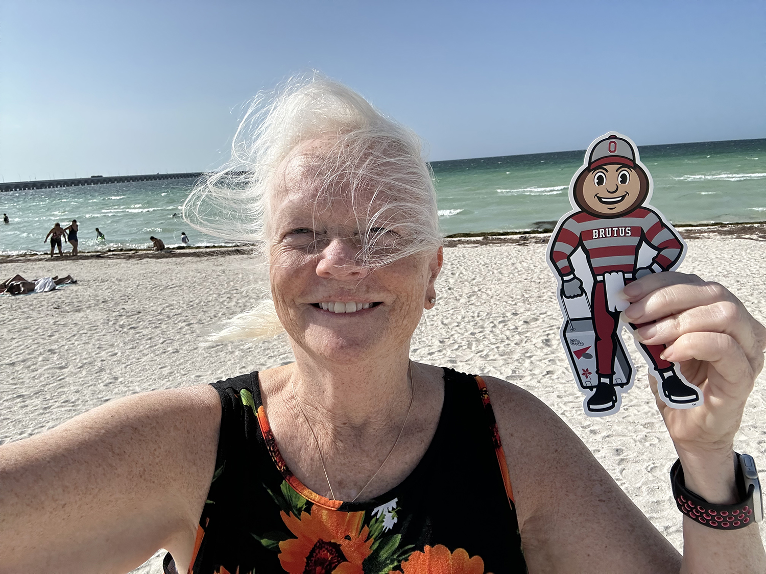 A woman smiling and holding Brutus on a white sandy beach with blue ocean water and people swimming in the background in Progreso, Mexico.