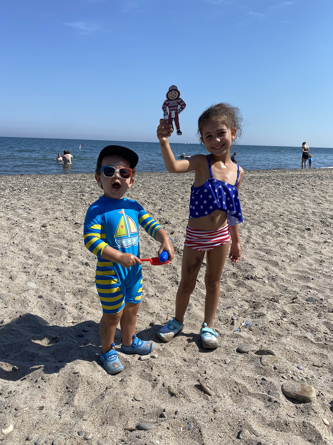 A little boy holding a shovel and a little girl holding Brutus, both wearing bathing suits, on a beach with Lake Erie and people swimming in the background in Geneva-on-the-Lake, Ohio.
