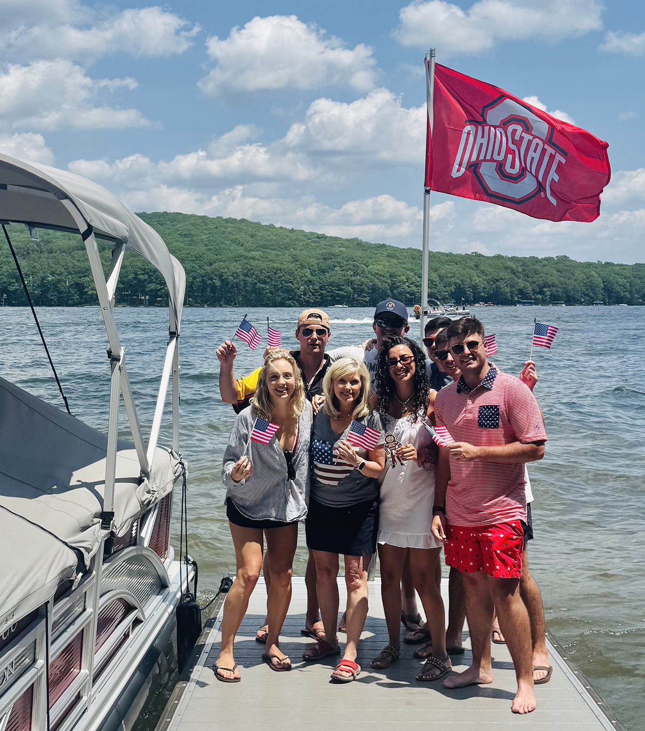 Three women and five men together on a dock by a boat with a lake behind them. One woman is holding Brutus while the others hold American flags, and an Ohio State flag flies behind them.