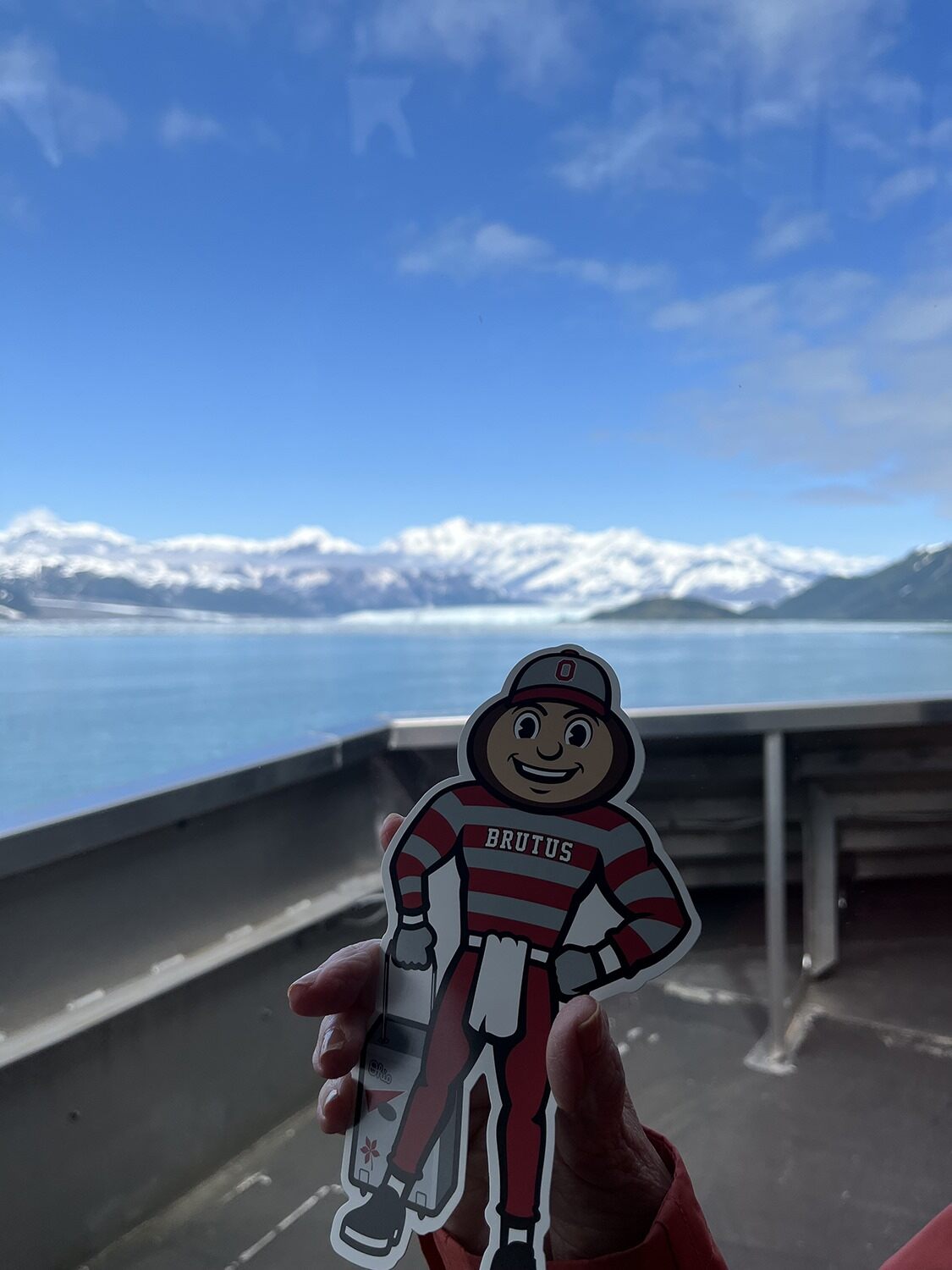 Brutus Buckeye cutout held in front of snow-capped mountains and lake in Hubbard Glacier, Alaska.