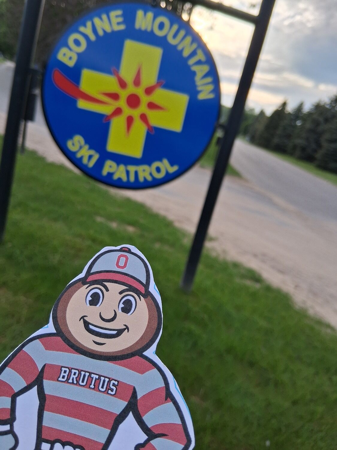 Brutus Buckeye cutout in front of a Boyne Mountain Ski Patrol sign with trees and a road in the background.