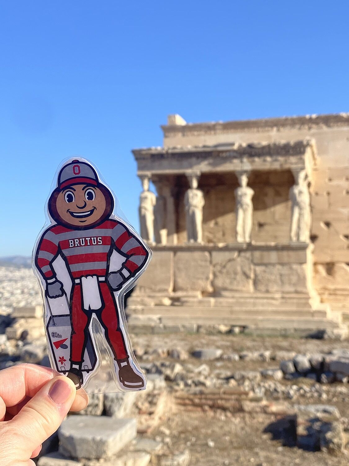Hand holding Brutus Buckeye cutout in front of ancient Greek temple with female statues as columns.