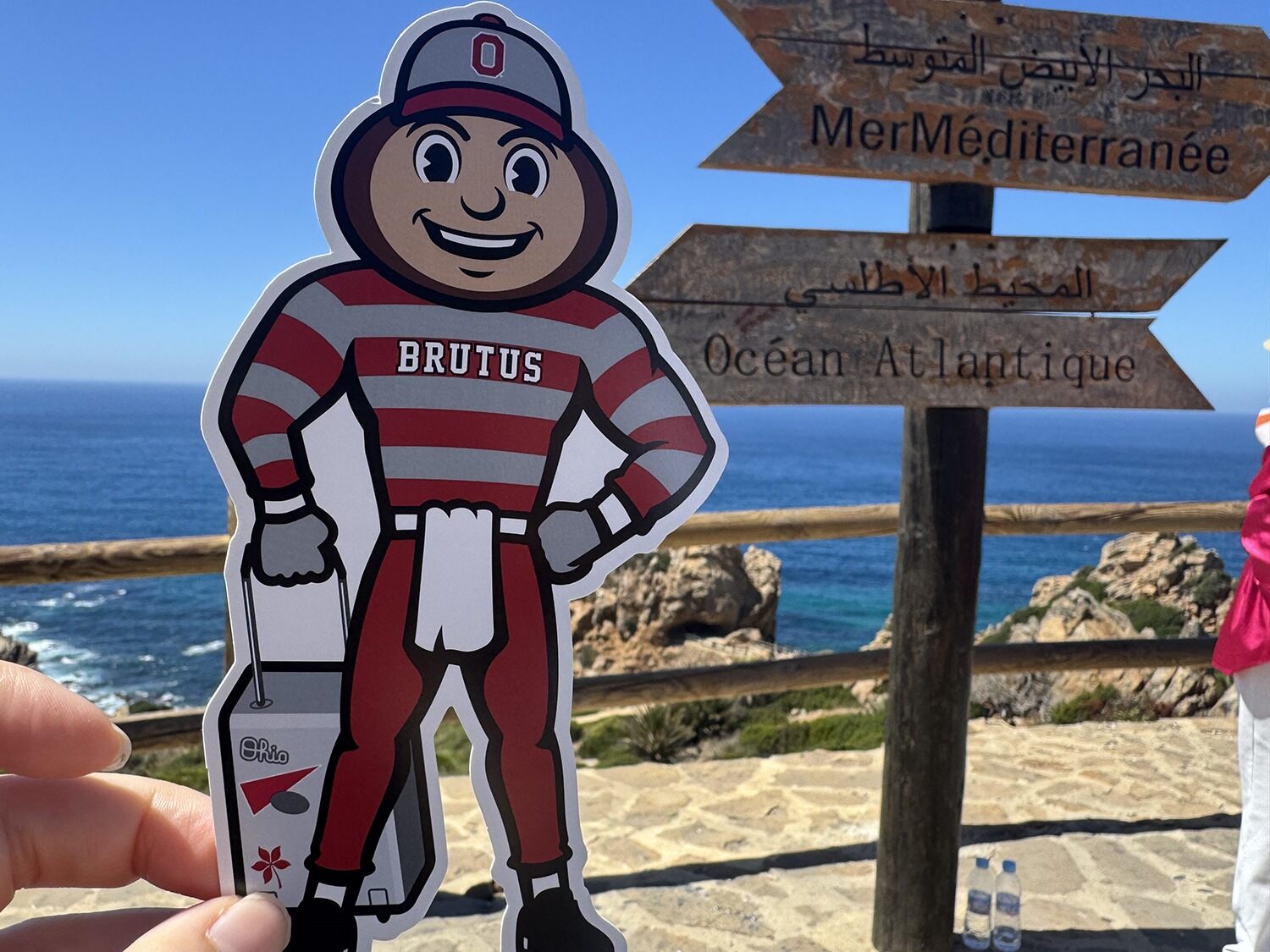 Brutus Buckeye cutout held in front of signposts for the Mediterranean Sea and Atlantic Ocean, with a coastal view in the background.