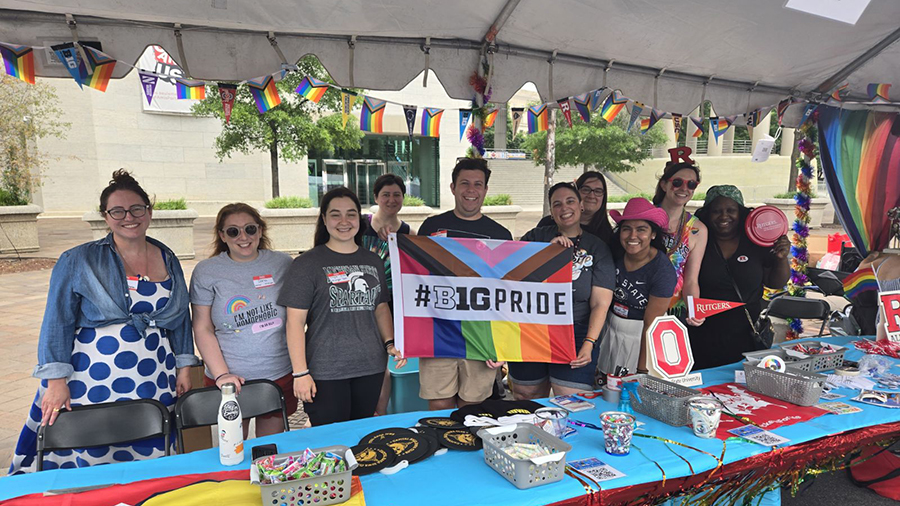A group of people stand under a tent decorated with rainbow Pride flags, holding a colorful Pride banner that reads “#B1GPRIDE.” A table in front of them is covered with bright cloths and various giveaway items.