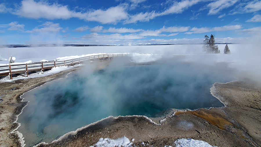 A steaming hot spring with deep blue water surrounded by snow under a bright, partly cloudy sky.