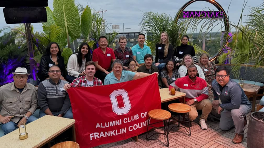 A group of people gathered on an outdoor rooftop holding a red flag that reads “Alumni Club of Franklin County,” surrounded by plants and patio seating.