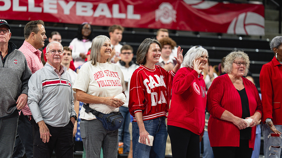 People standing in a sports arena wearing Ohio State apparel, with a red “Volleyball” banner visible in the background.