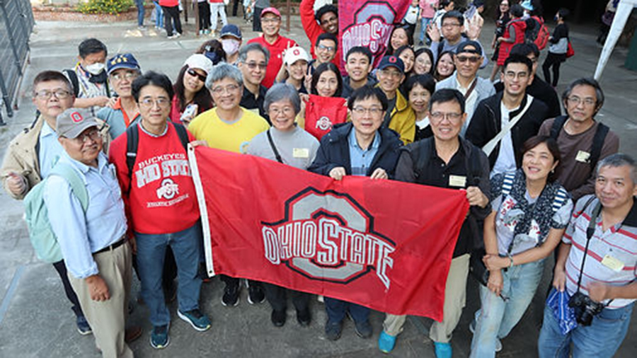A large group of people gathered outdoors holding a red Ohio State flag with the university’s logo, standing closely together for a group photo.