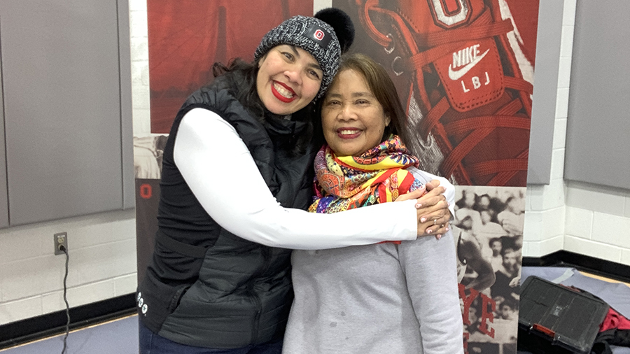 Two people standing close together indoors, with one person’s arm around the other. They are in front of a display featuring sports-themed graphics, including a large red shoe with a Nike logo.