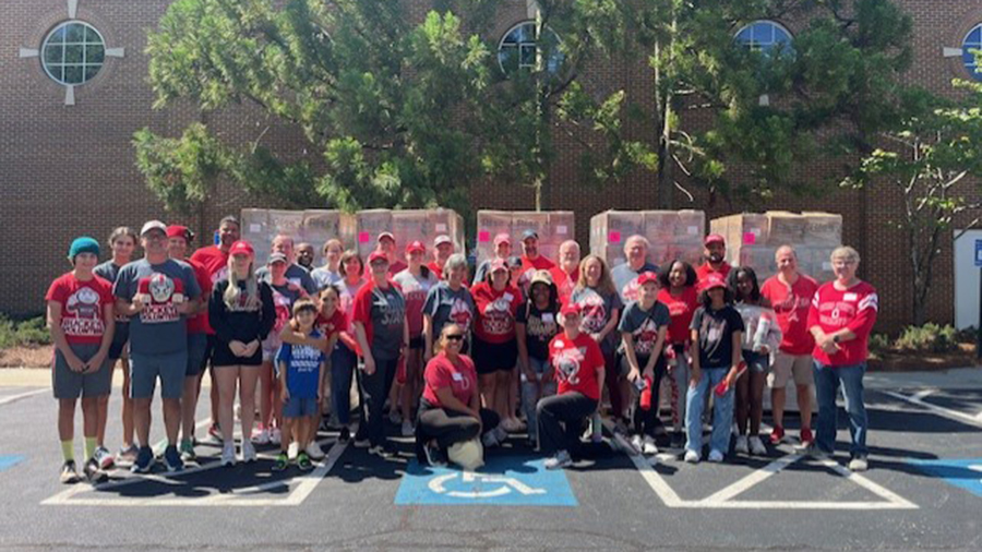 A large group of people wearing coordinated red, gray, and black clothing stand together outdoors in a parking lot. They are positioned in front of several stacked pallets wrapped in plastic, with a brick building and tall trees in the background.