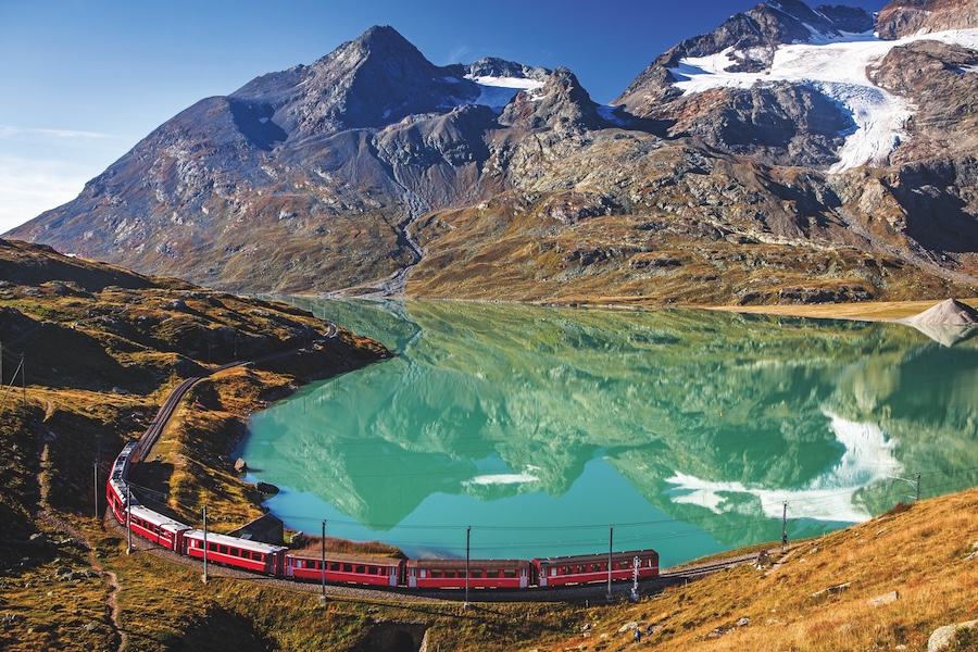 A train traveling past a greenish blue lake and snow-capped mountains in the distance