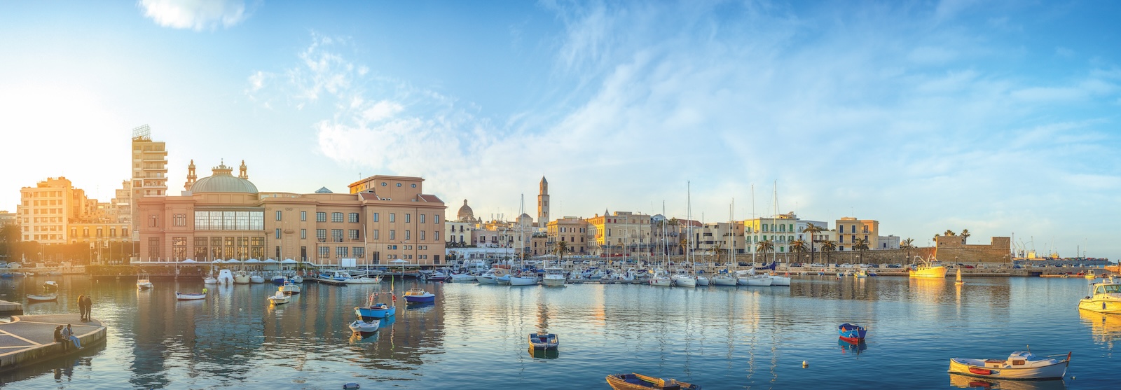 Harbor view of Bari with fishing boats, waterfront buildings, and calm sea at sunset.