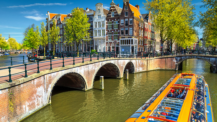 A canal boat passes under a brick bridge in Amsterdam, with traditional narrow houses and trees lining the water on a sunny day.