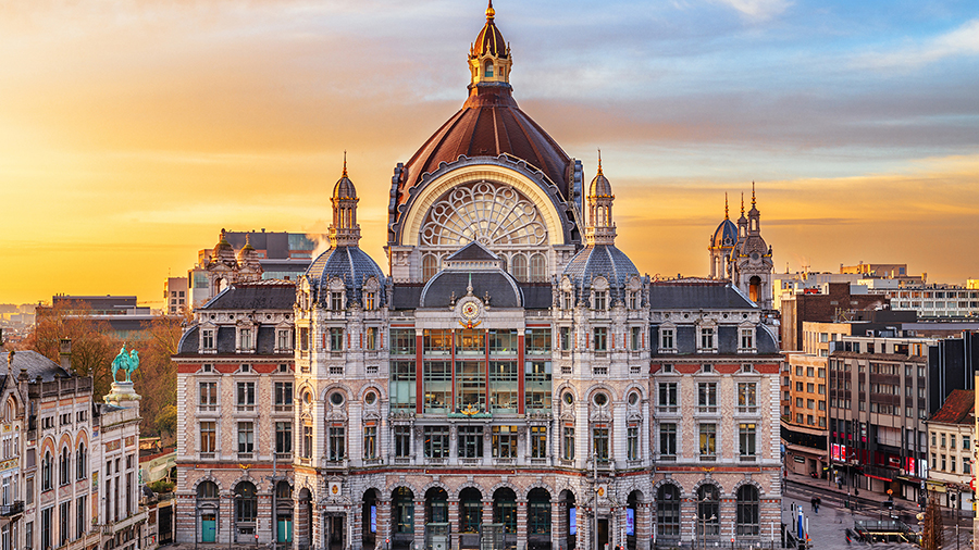 A grand, ornate building with a large domed roof is illuminated by a golden sunset. The foreground shows an empty plaza, flanked by city architecture.