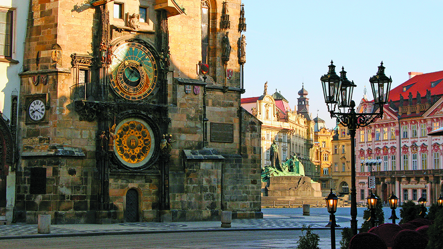 An ornate astronomical clock on the side of a historic building in a European square, with nearby lampposts and surrounding colorful architecture.