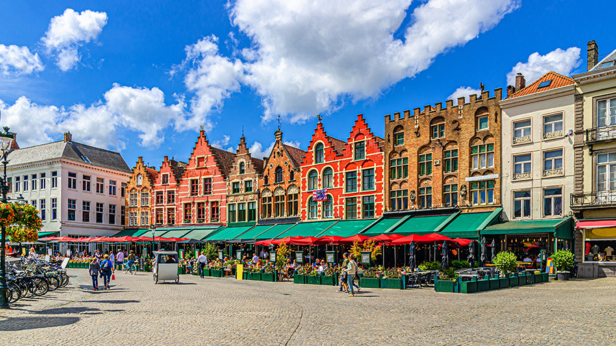 Vibrant market square with colorful, historic gabled buildings under a blue sky. People stroll and dine outdoors, creating a lively, cheerful atmosphere.