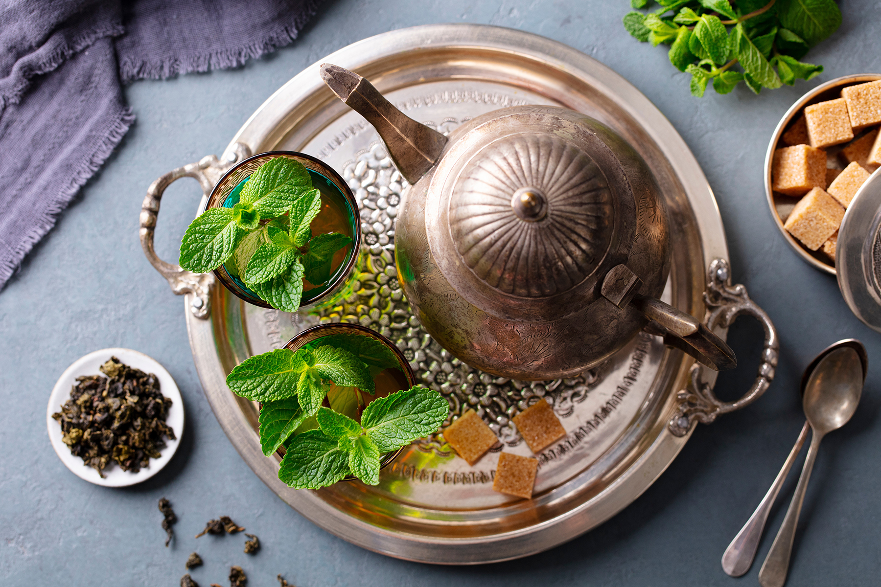 A silver tray holds a vintage teapot and two glasses of mint tea, garnished with fresh mint leaves. Sugar cubes, loose tea, and spoons surround the tray. Cozy and inviting.