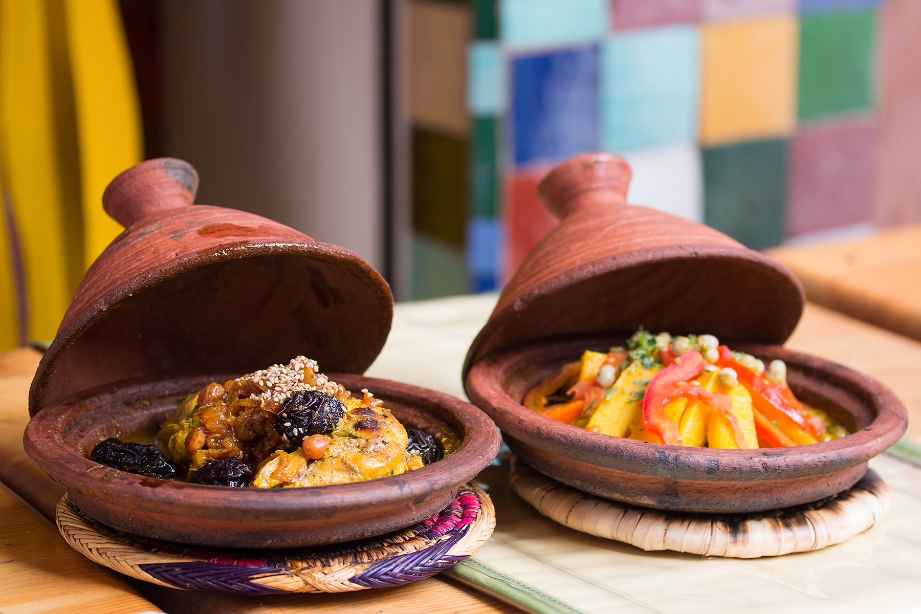 Two clay tagines filled with vibrant, colorful dishes. The left has a savory stew garnished with nuts and dried fruits; the right features vegetables and chickpeas. A checkered wall backdrop adds warmth.