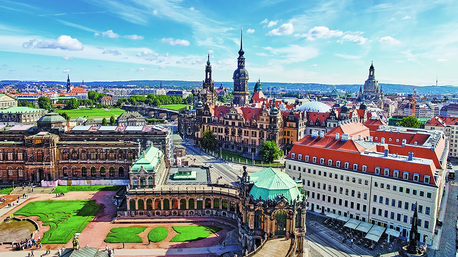 An aerial view of Dresden city with ornate buildings, towers, and red-roofed structures under a bright blue sky.