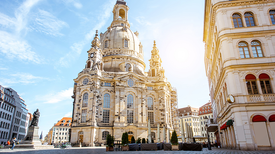 A large ornate stone church with a tall dome and detailed façade, standing in a bright open square on a sunny day.