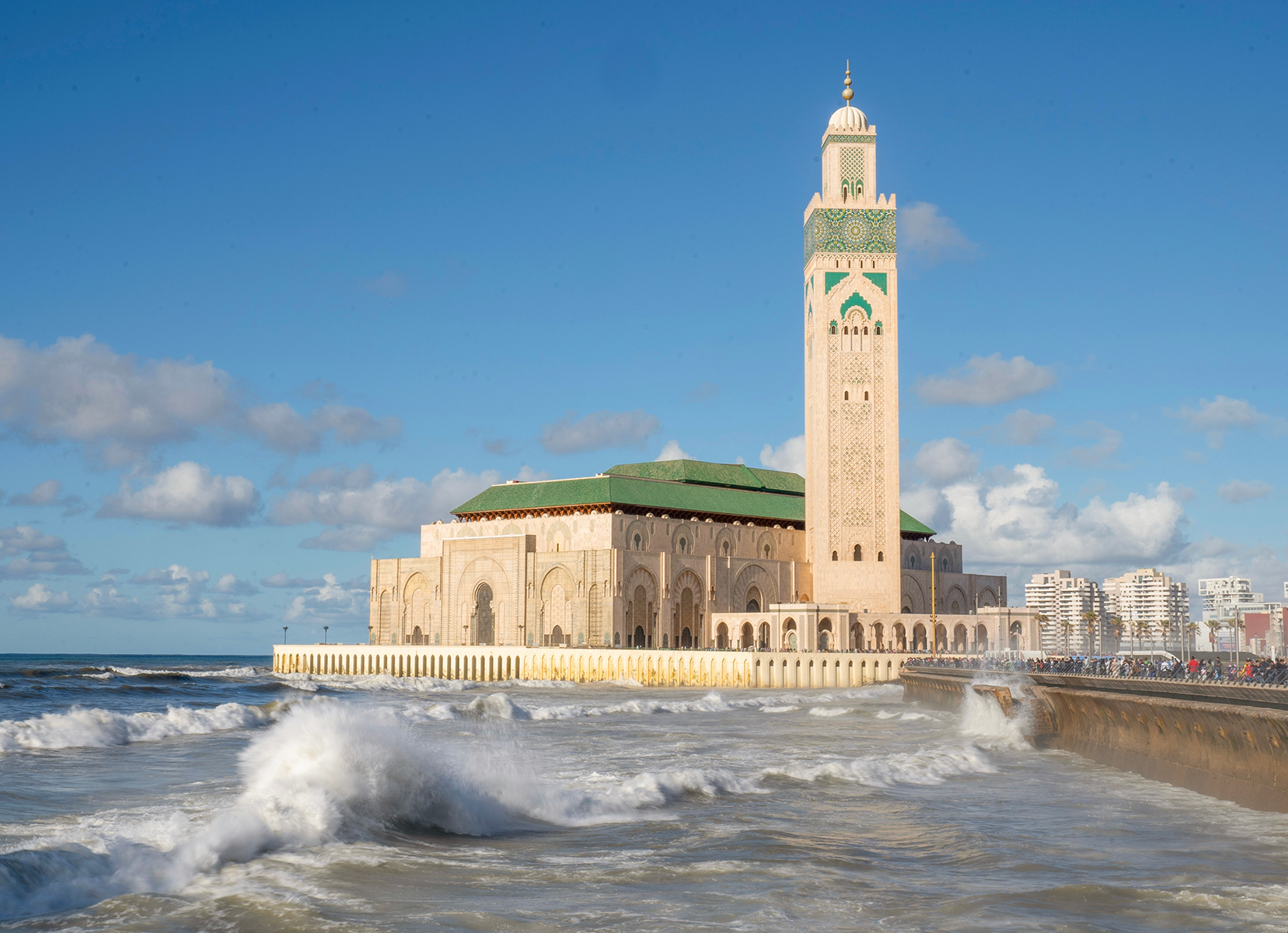 The image shows the Hassan II Mosque in Casablanca, Morocco, with its tall minaret and green roof, set against a blue sky. Waves crash against the shore.