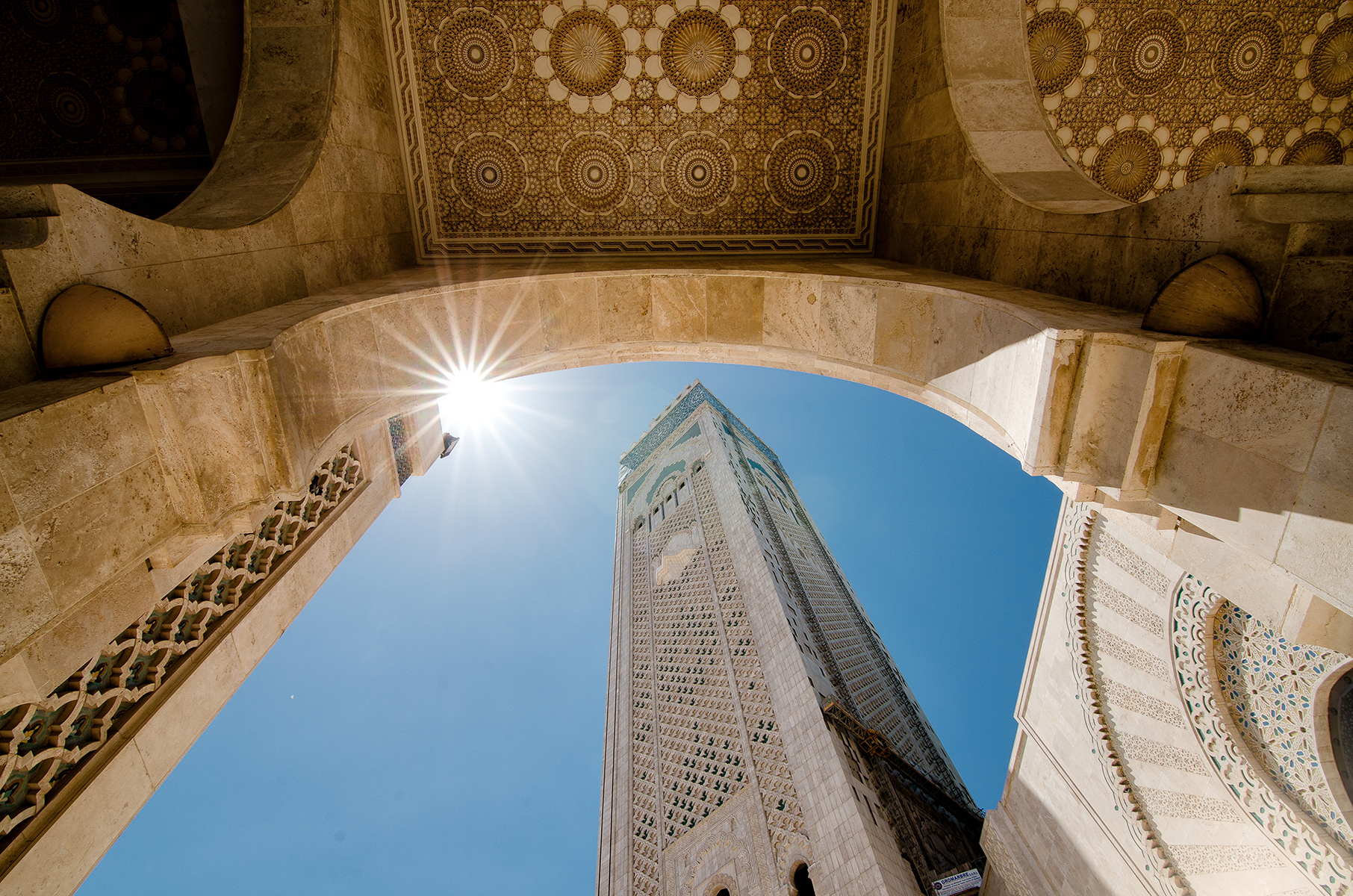 Tall minaret viewed from below through an ornate archway against a clear blue sky. The sun peeks over the arch, casting rays and evoking a serene atmosphere.