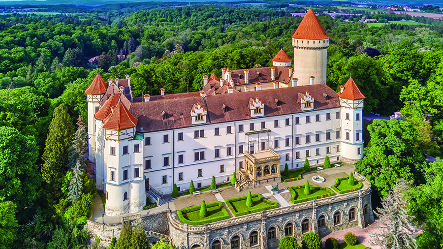An aerial view of a Konopiste Castle with red-roofed towers surrounded by dense green forest.