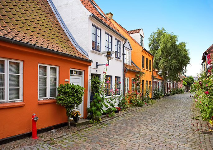 Colorful houses along a cobblestone street in Aarhus with flowers and greenery.