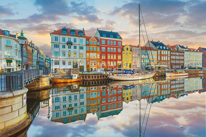 Colorful buildings in Copenhagen reflected in a calm canal with boats along the waterfront.