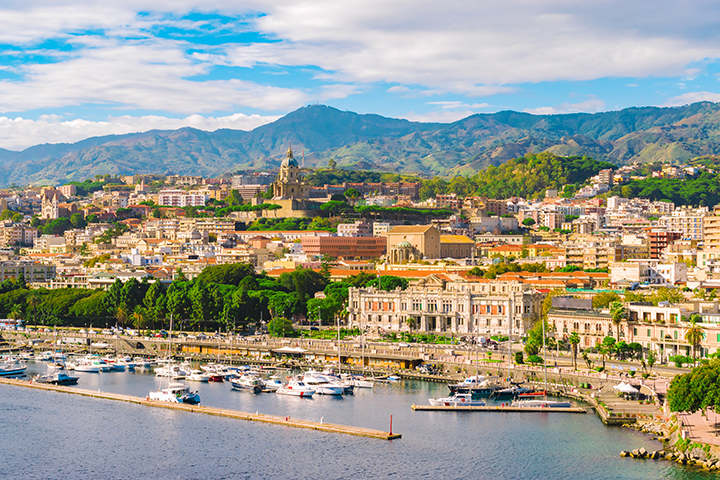 Messina waterfront with marina, historic buildings, and mountain backdrop.