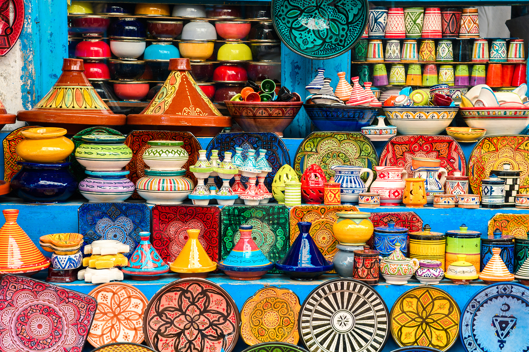 A vibrant display of colorful Moroccan pottery, including plates, bowls, and tagines, arranged on shelves. Patterns and vivid hues evoke a lively, cultural atmosphere.