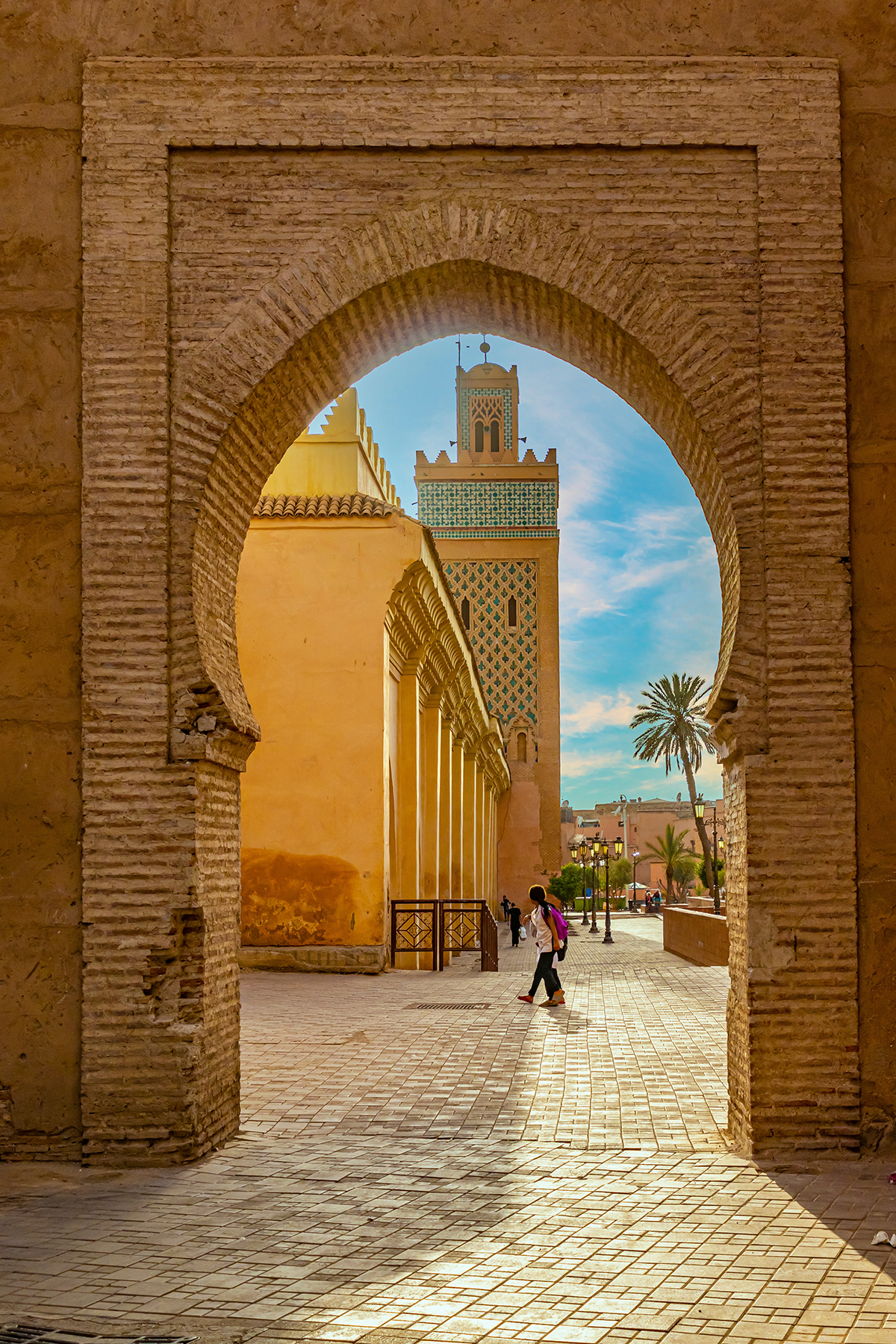 A stone arch frames a sunlit courtyard with a towering mosque featuring intricate patterns. A person walks under the arch, conveying a sense of calm.