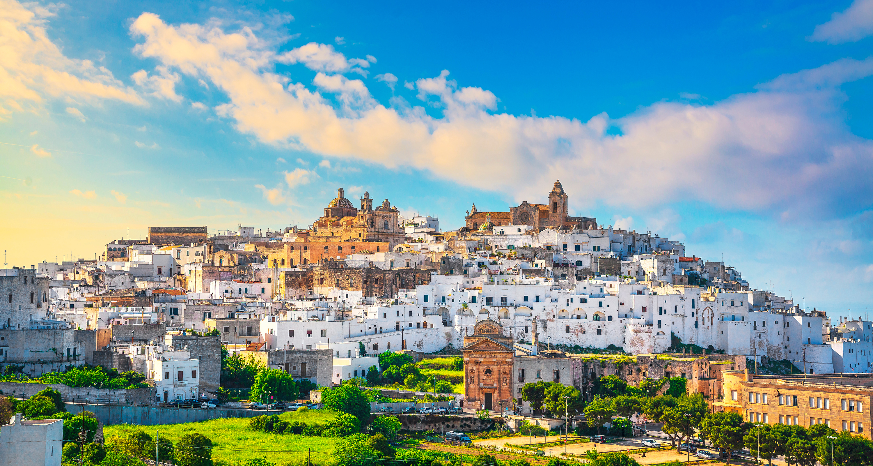 Sunlit hillside view of Ostuni’s whitewashed old town surrounded by green countryside.