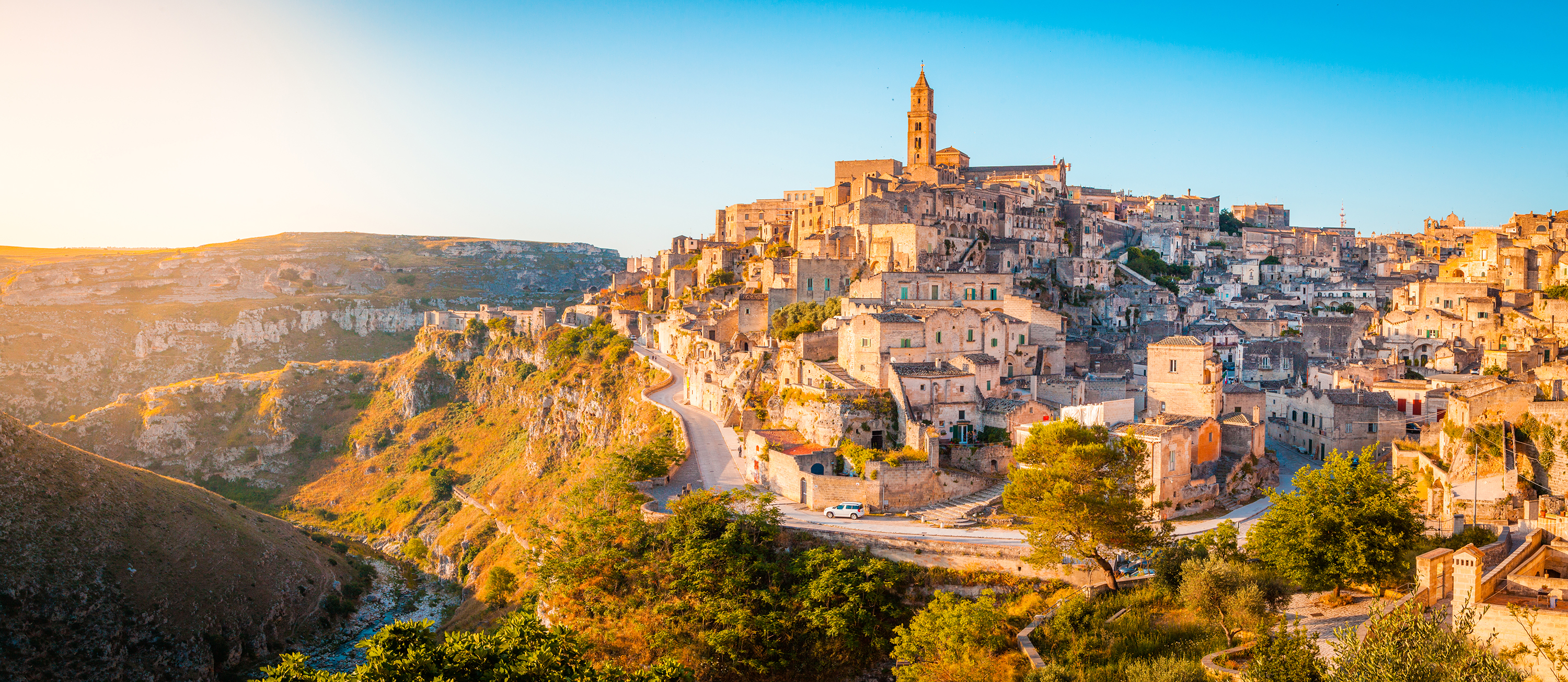 Panoramic view of the ancient hilltop city of Matera with stone buildings and winding roads at sunset.